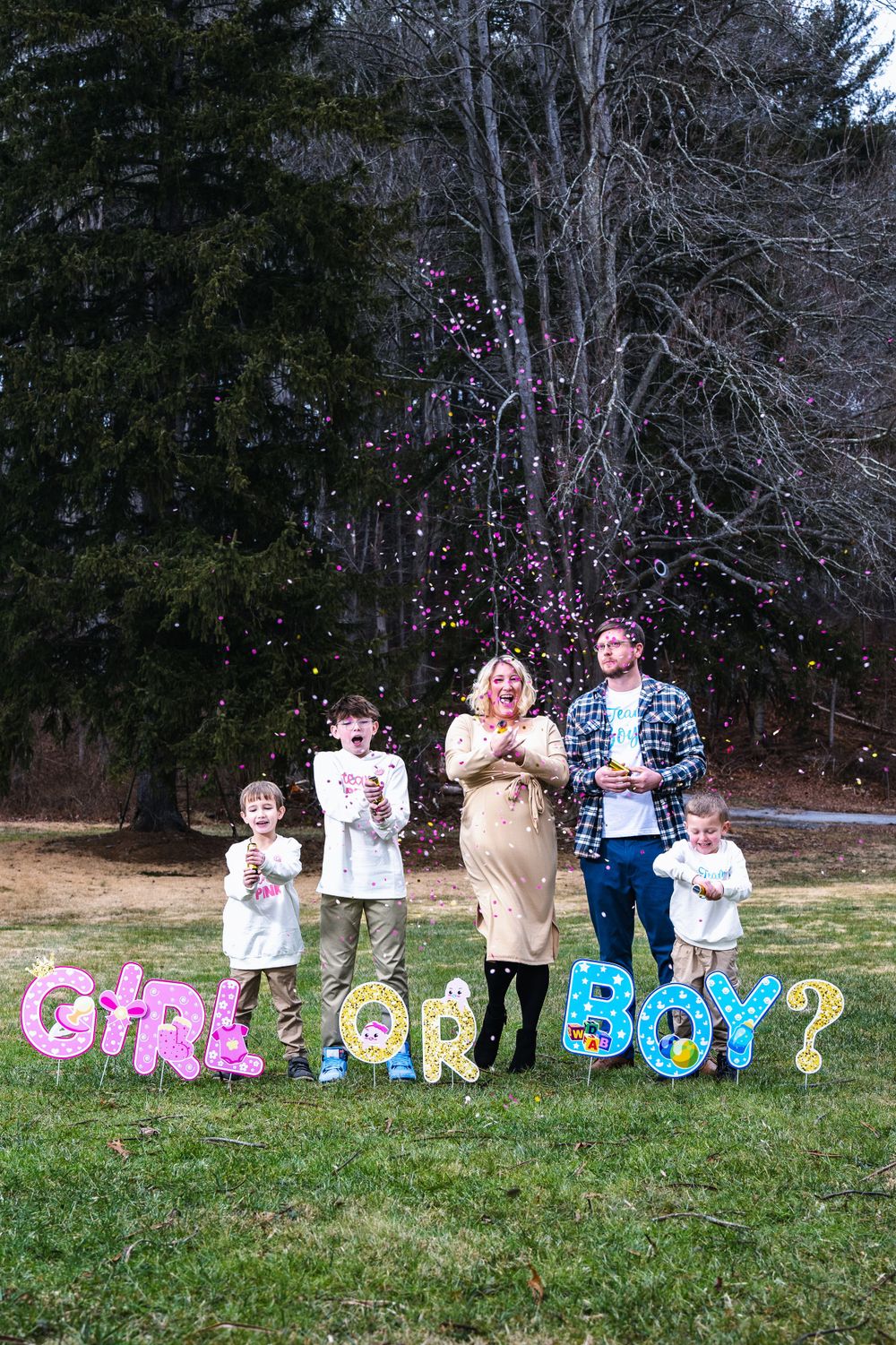 A gender reveal celebration outdoors with pink and blue balloon letters spelling 'GIRL OR BOY' on a grassy lawn.