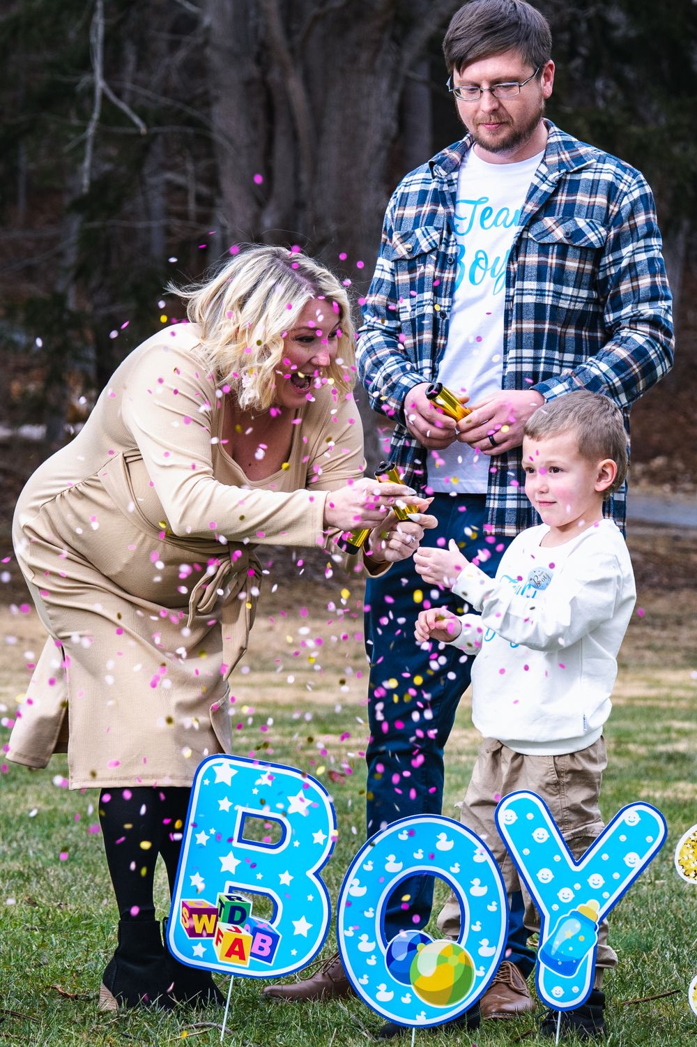 Family celebrates gender reveal with blue confetti and letters spelling BOY during outdoor announcement photoshoot.