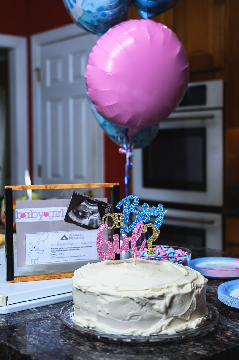 Baby shower celebration setup with white frosted cake, pink and blue balloons, and decorative cake topper on dark counter.