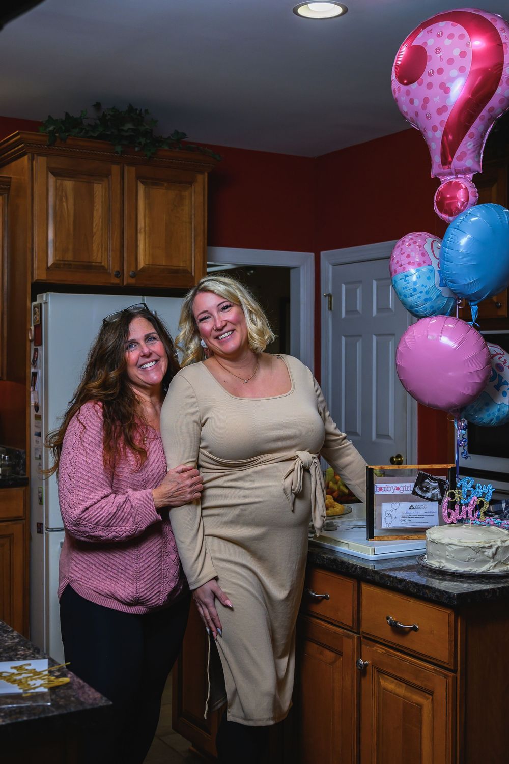 Two women celebrating in a kitchen with colorful balloons including a pink question mark balloon decoration.