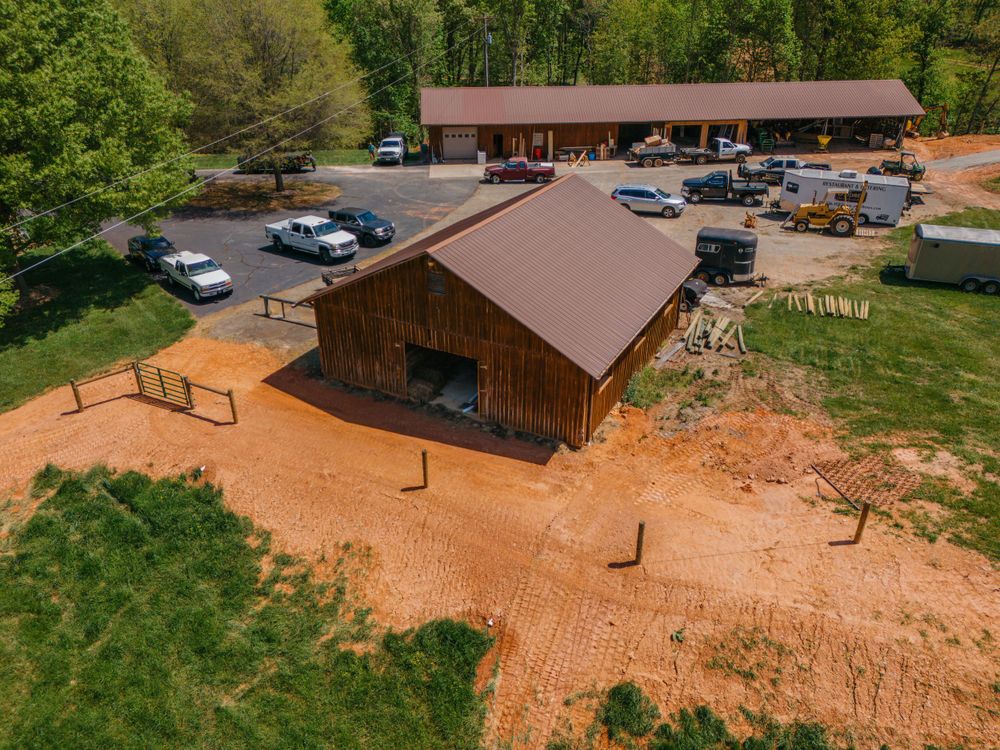 Aerial view of a rustic wooden barn structure with brown metal roof and dirt parking area surrounded by trees.
