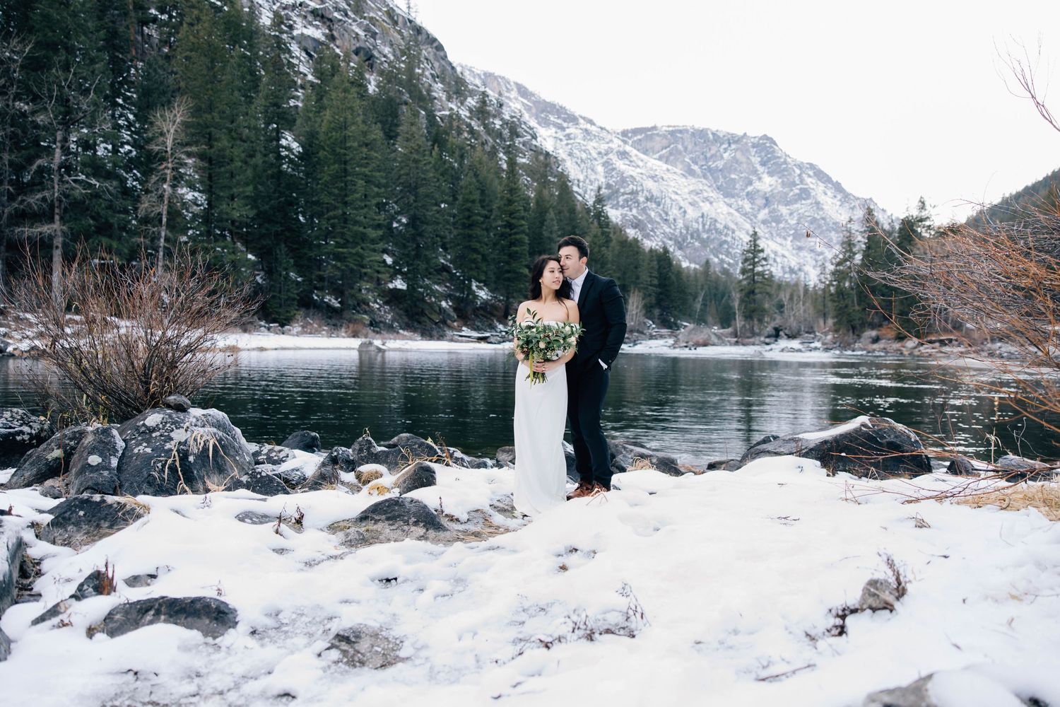 Newlywed couple embracing in snowy winter landscape beside a mountain lake surrounded by pine trees.