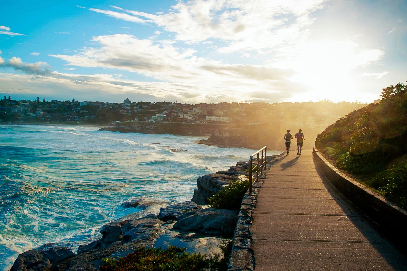 Coastal walkway at sunset with silhouetted figures walking along rocky oceanside path.