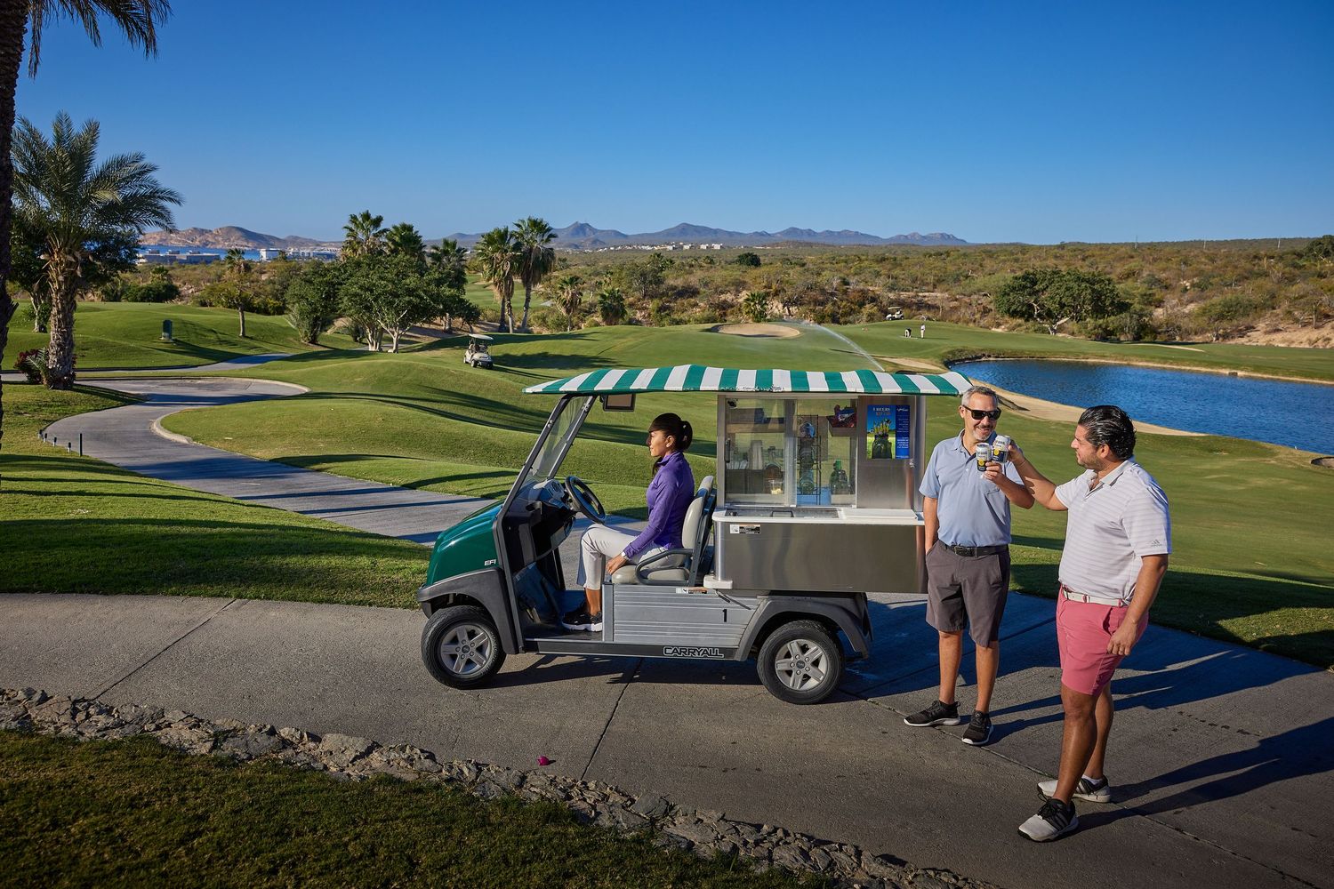 Group gathered around golf cart with striped awning on scenic golf course overlooking ocean.