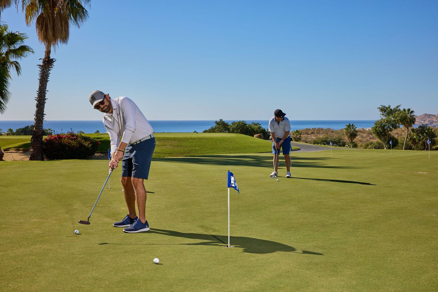 Golfers practicing putting on green with ocean and palm trees visible in background.