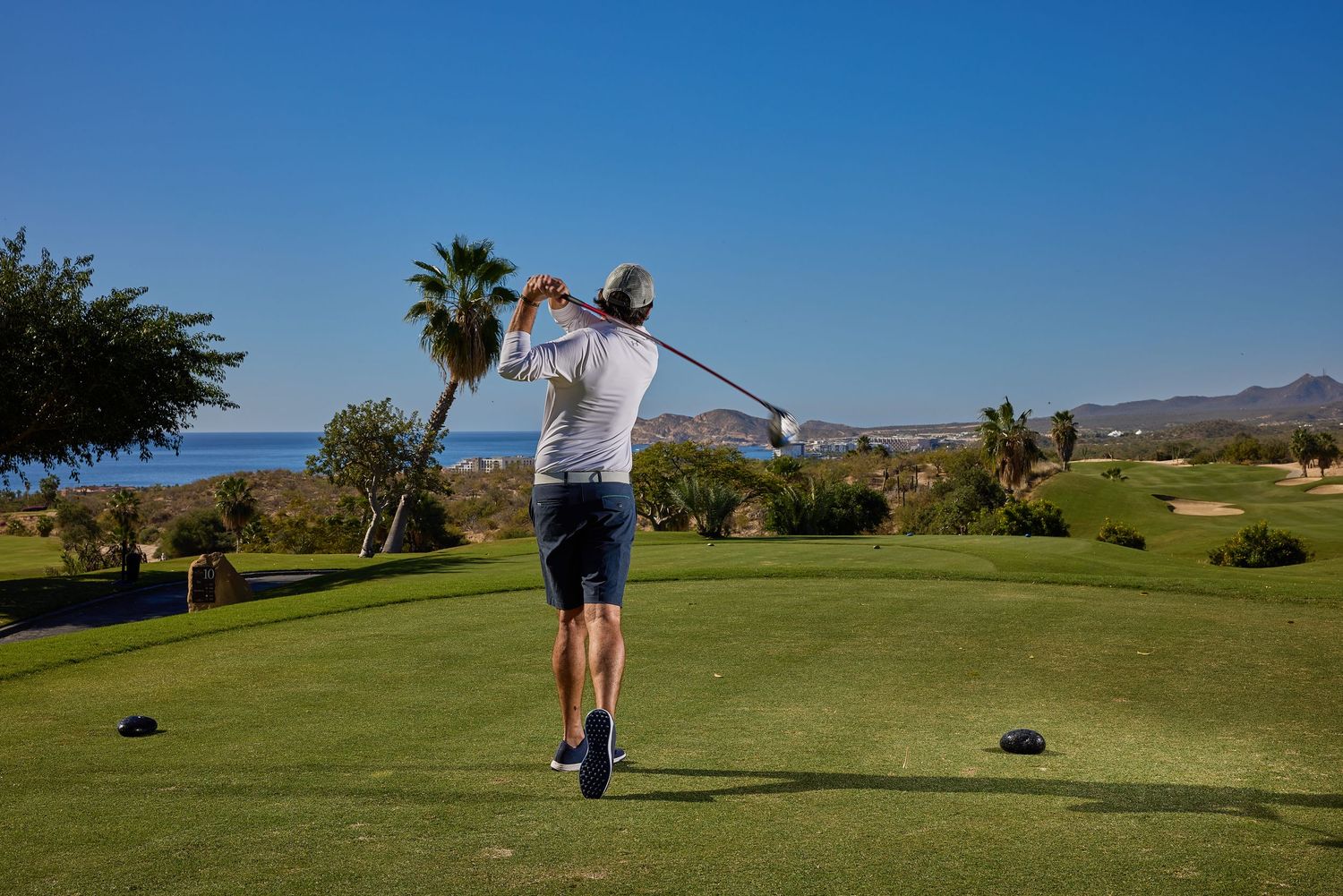 Golfer taking a swing on sunny golf course with palm trees and mountains in background.