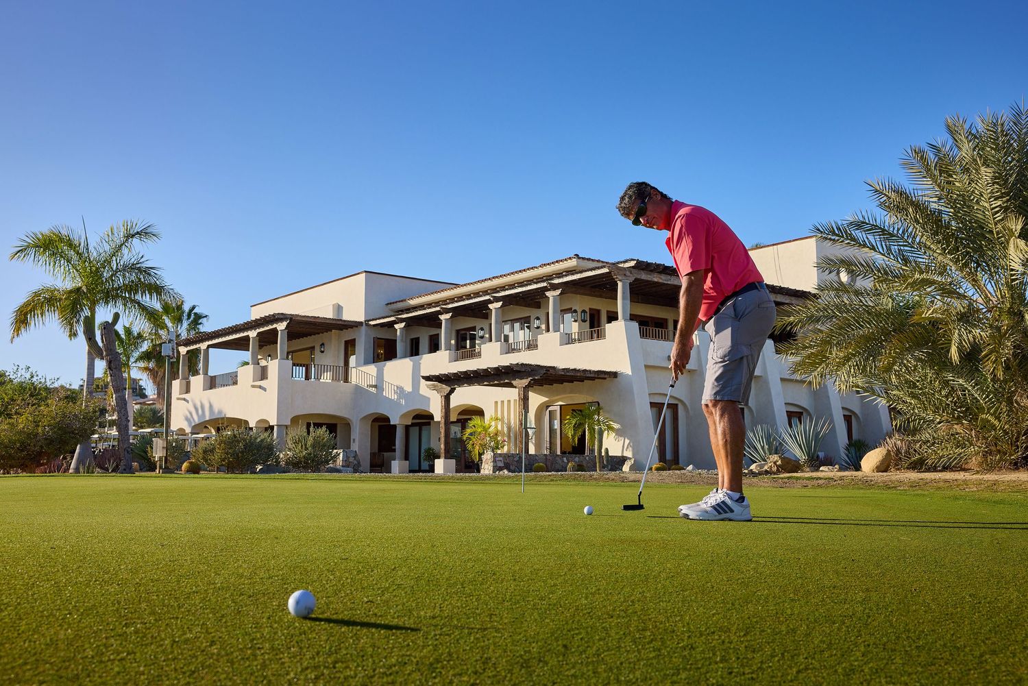 Golfer putting on a manicured green with luxury resort building in background.