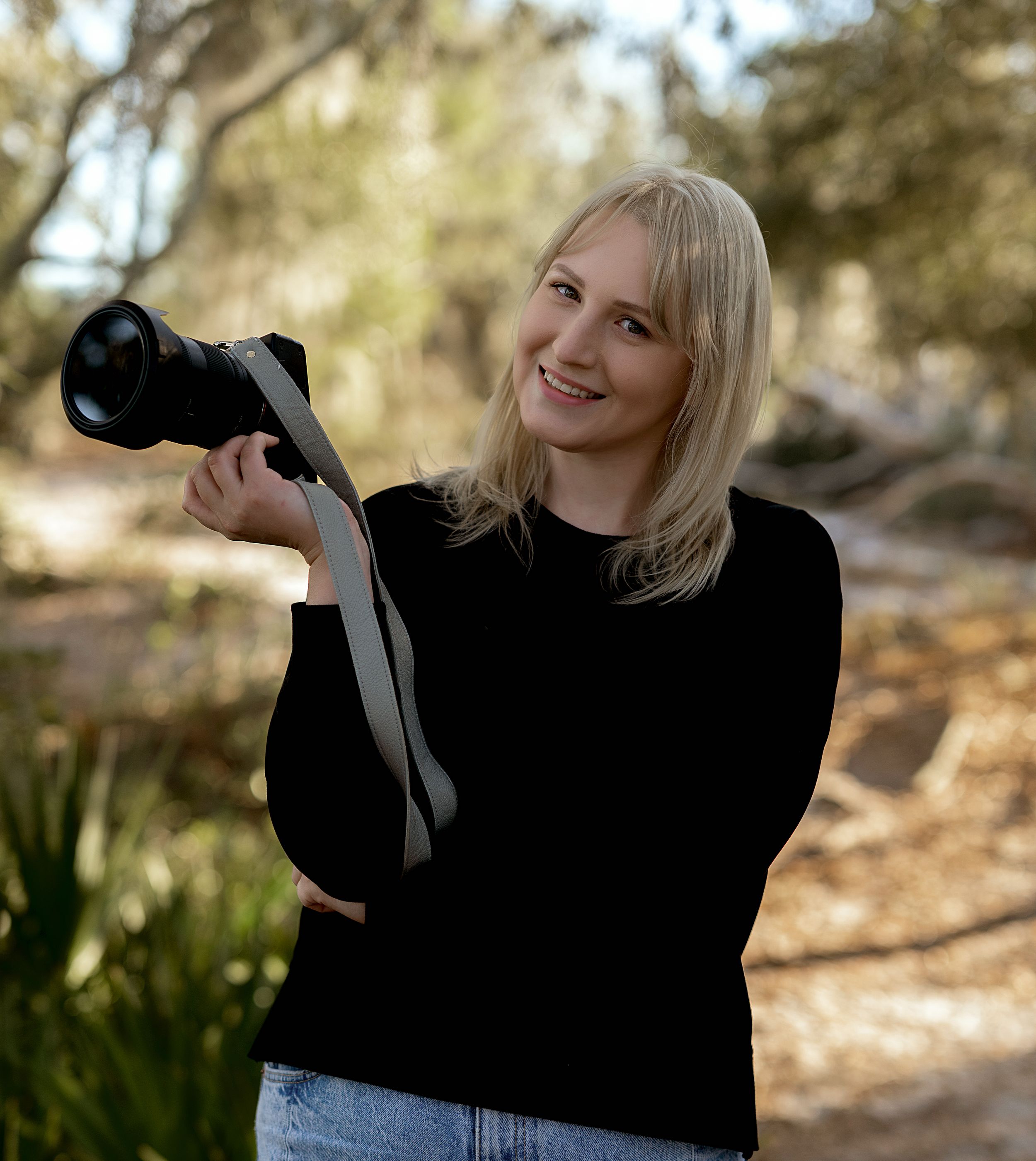 Photographer in black sweater holding professional camera outdoors in natural setting with blurred foliage background.