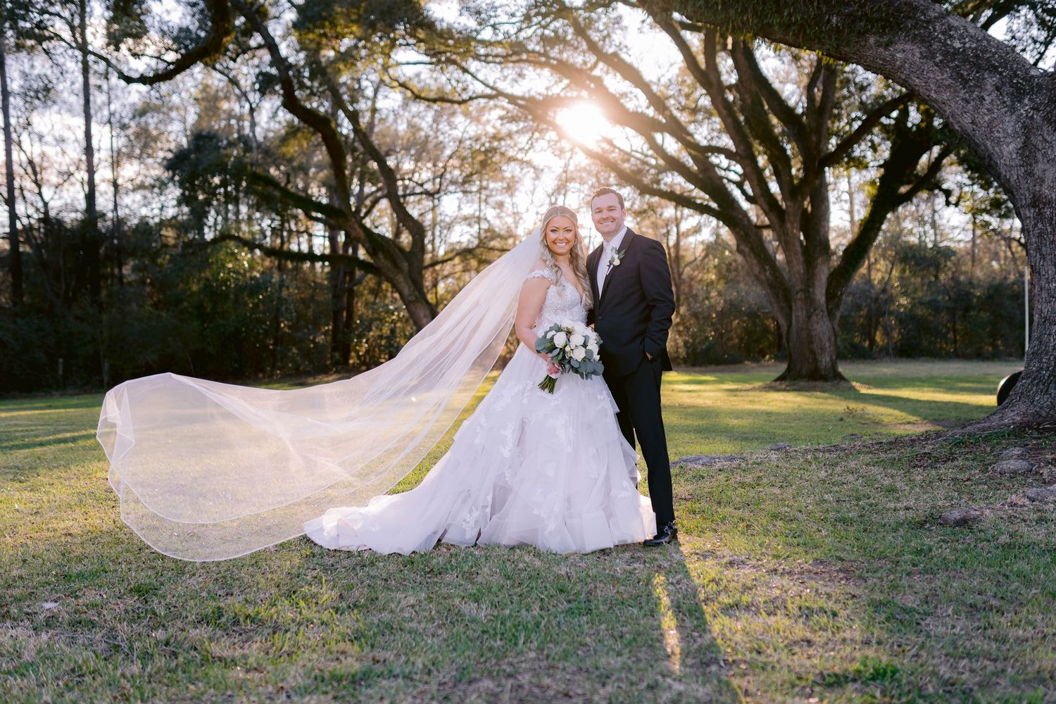 Wedding couple poses outdoors at sunset with bride's flowing veil catching the breeze under oak trees.