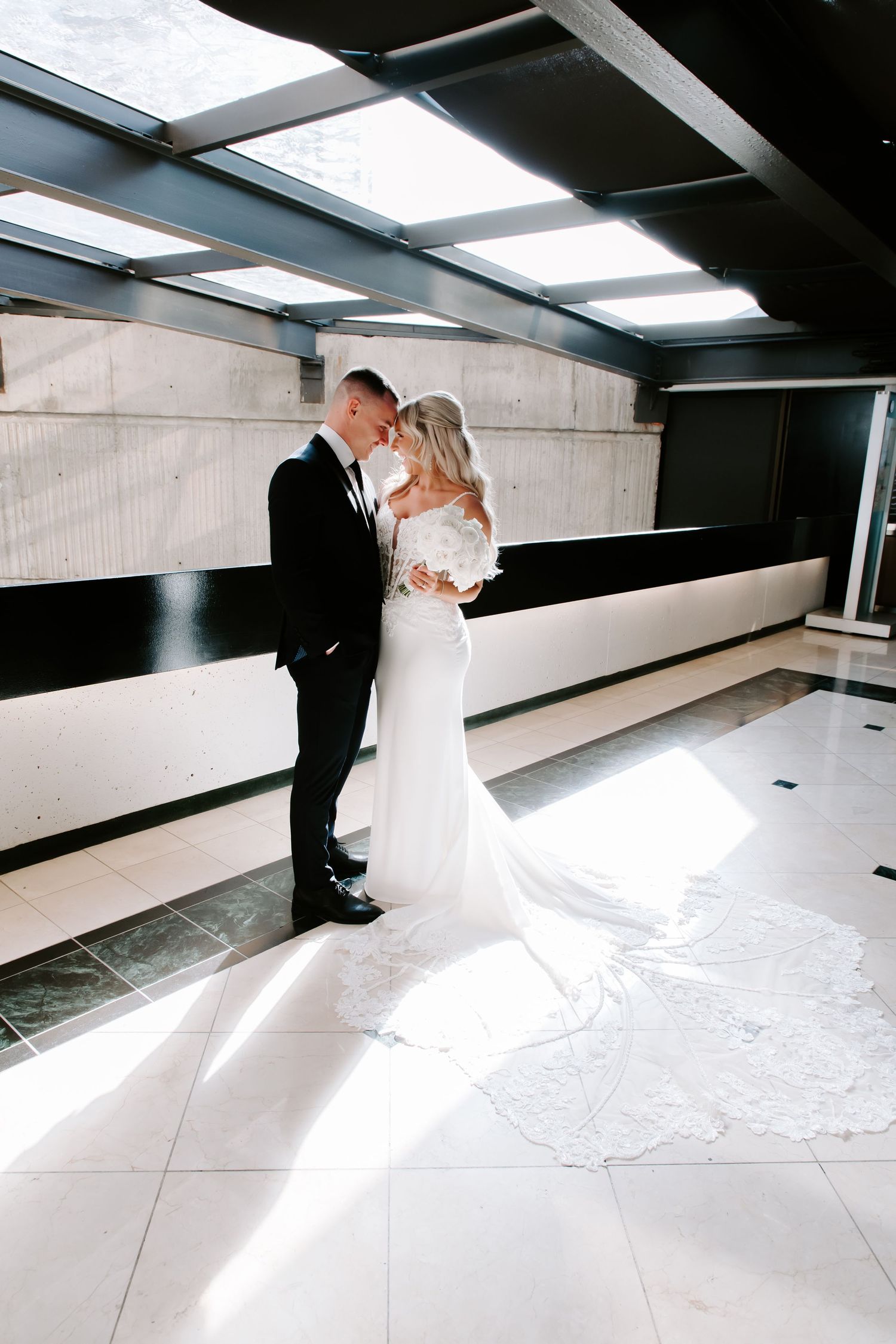 A romantic wedding couple embraces in a modern white interior space with dramatic lighting from skylights above.