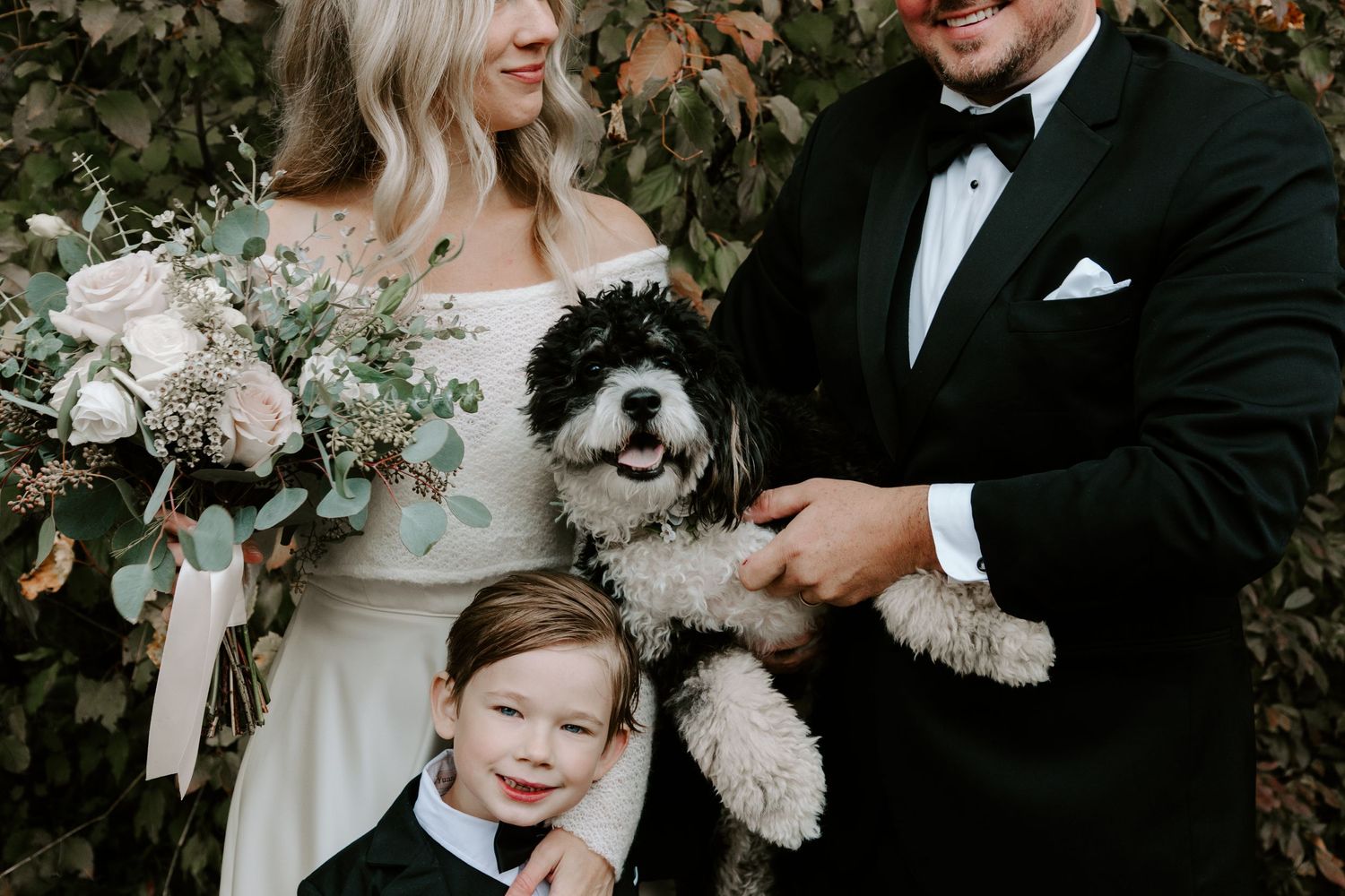 A wedding couple poses with a small child and fluffy white dog amidst autumn foliage.