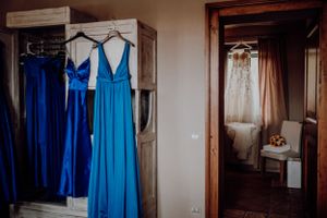 Blue bridesmaid dresses hanging on a wooden wardrobe with a white wedding gown visible in a mirror reflection.