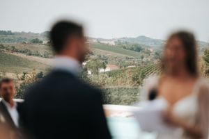Outdoor wedding ceremony with groom in navy suit and bride in white dress against scenic vineyard backdrop.