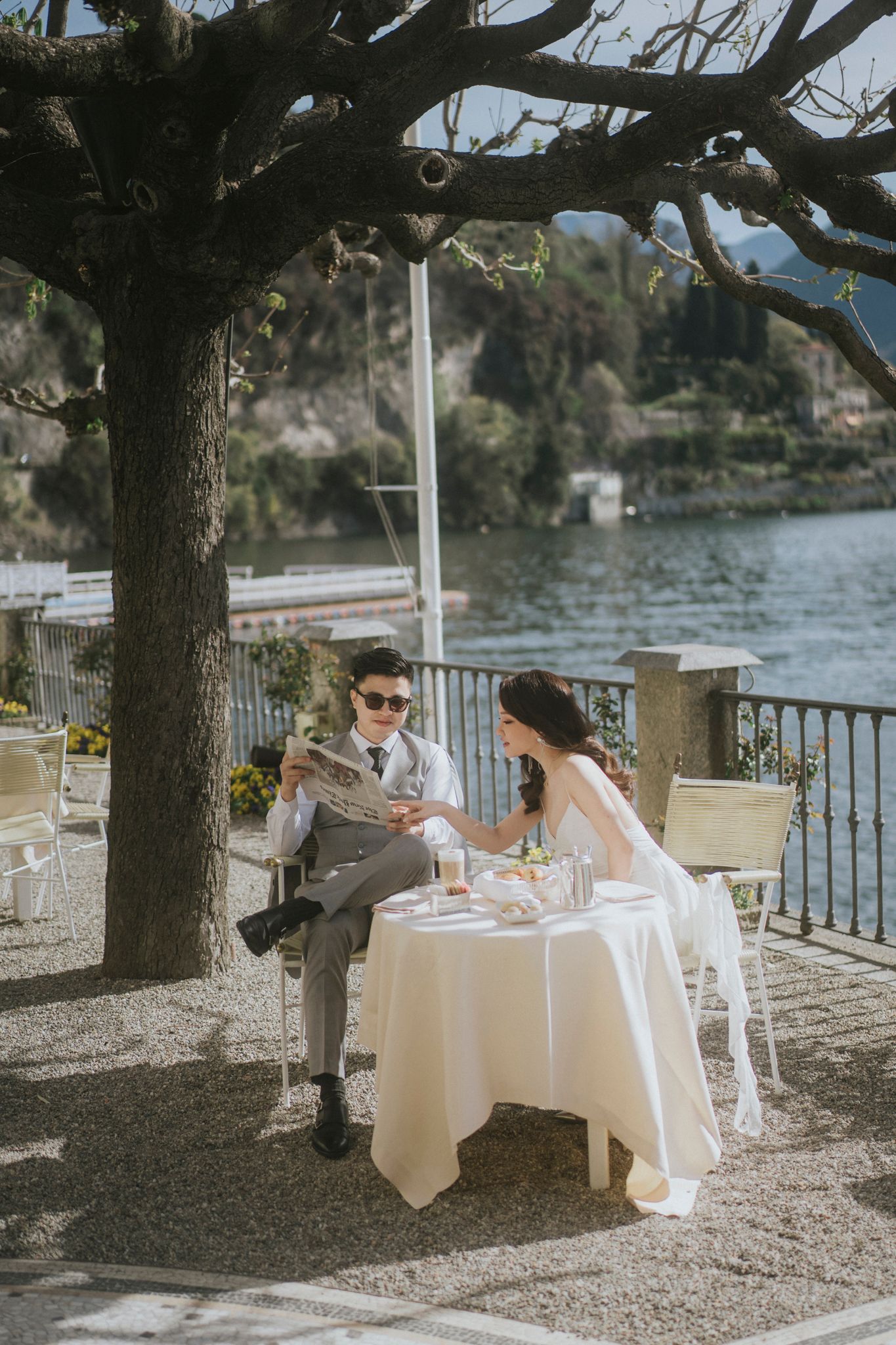 Elegant wedding couple dining at waterfront restaurant under large tree.