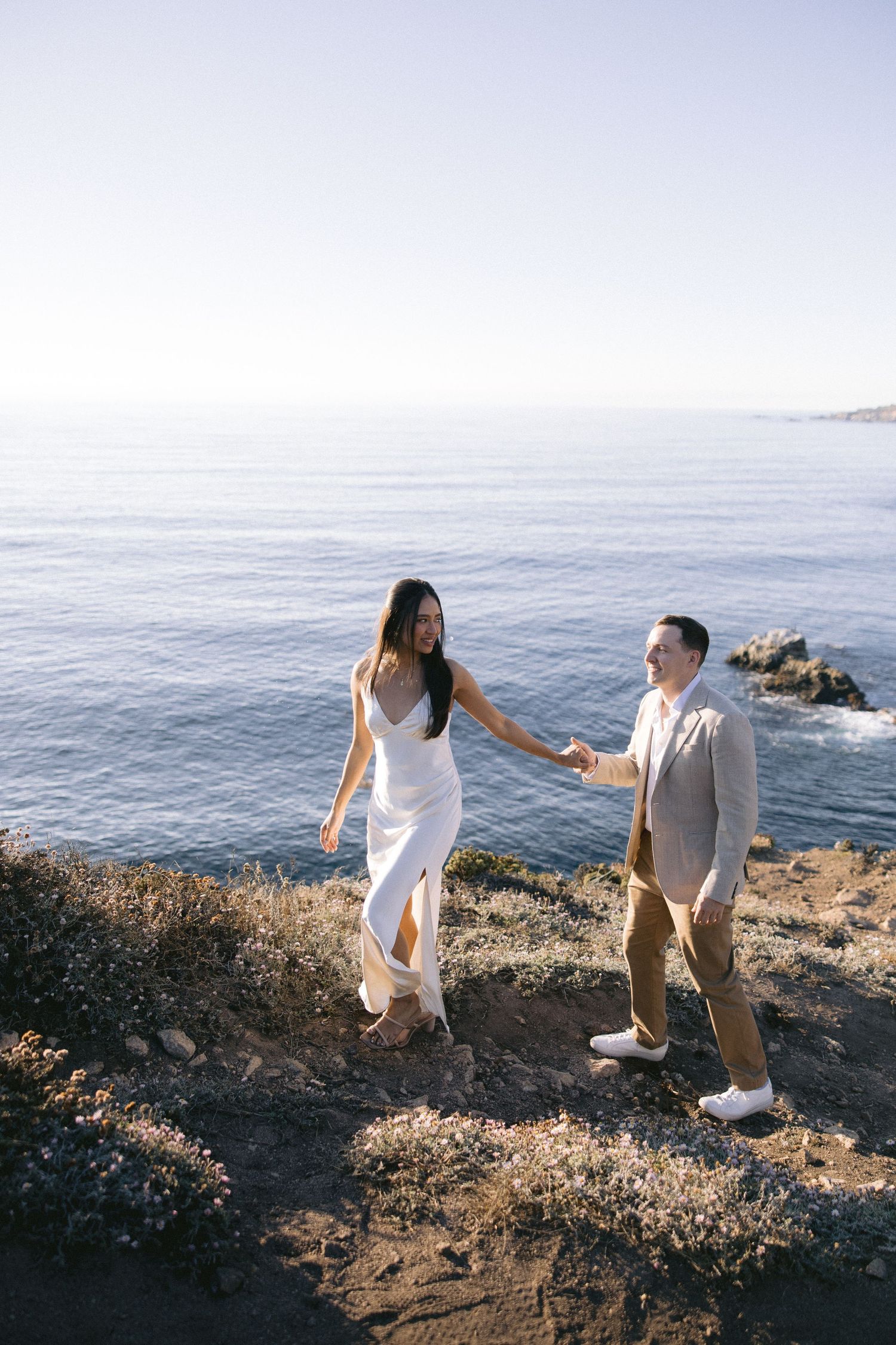 Couple holds hands while standing on coastal cliff overlooking blue ocean waters during romantic photoshoot.