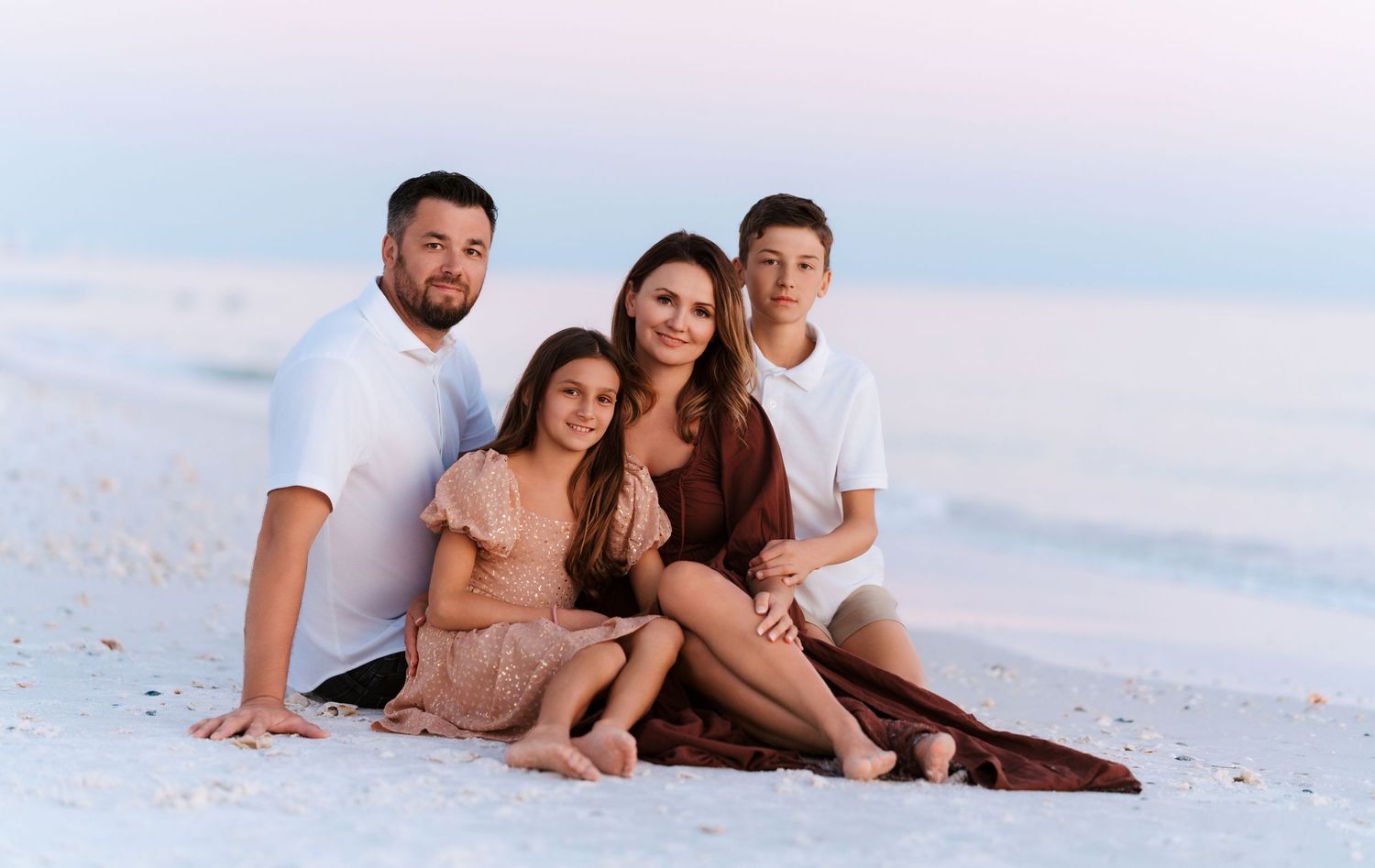 A family group sits together on a white sand beach wearing coordinated neutral-colored outfits at sunset.