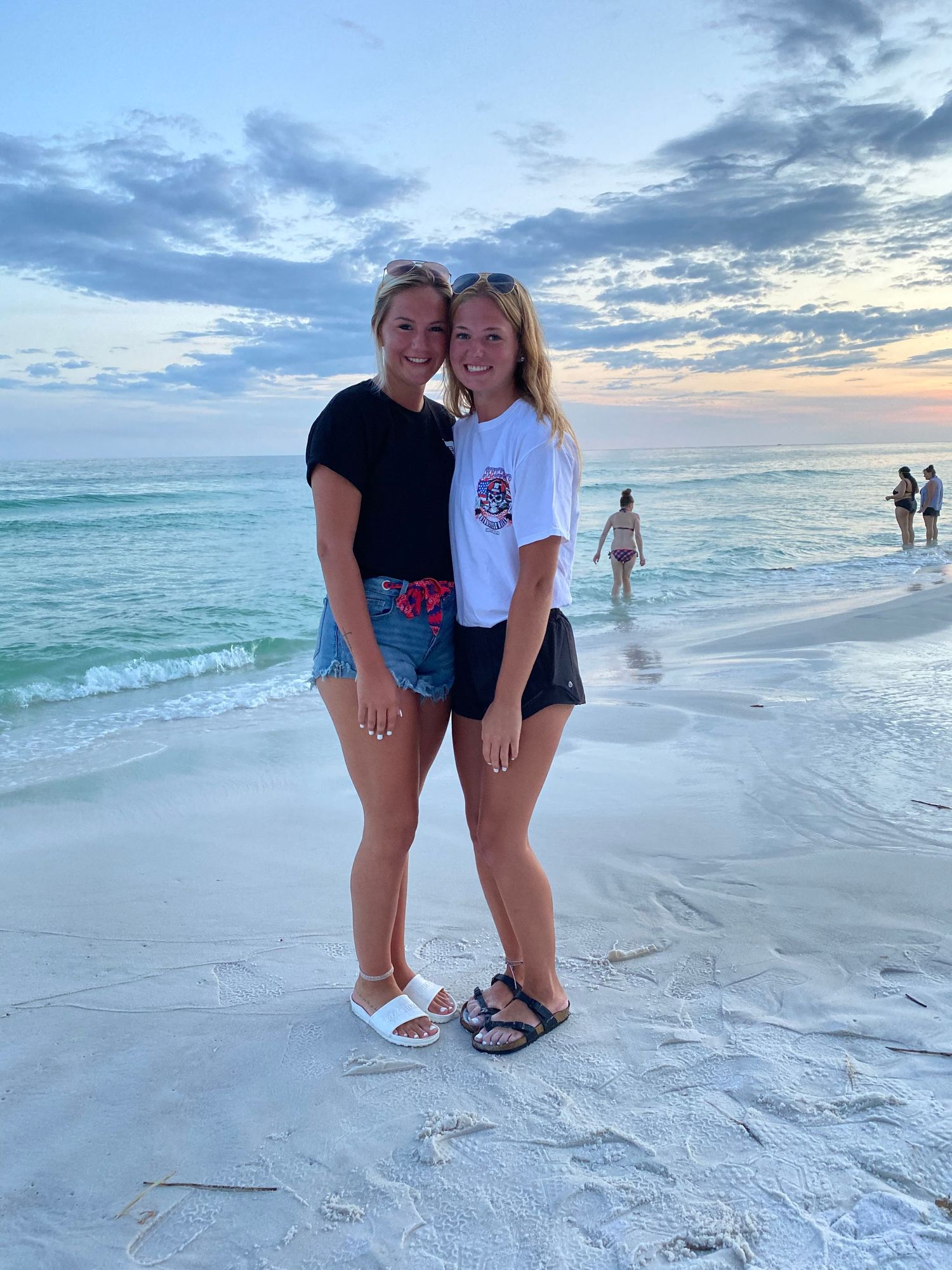 Two people pose together on a beach at sunset with turquoise waters and cloudy skies in the background.