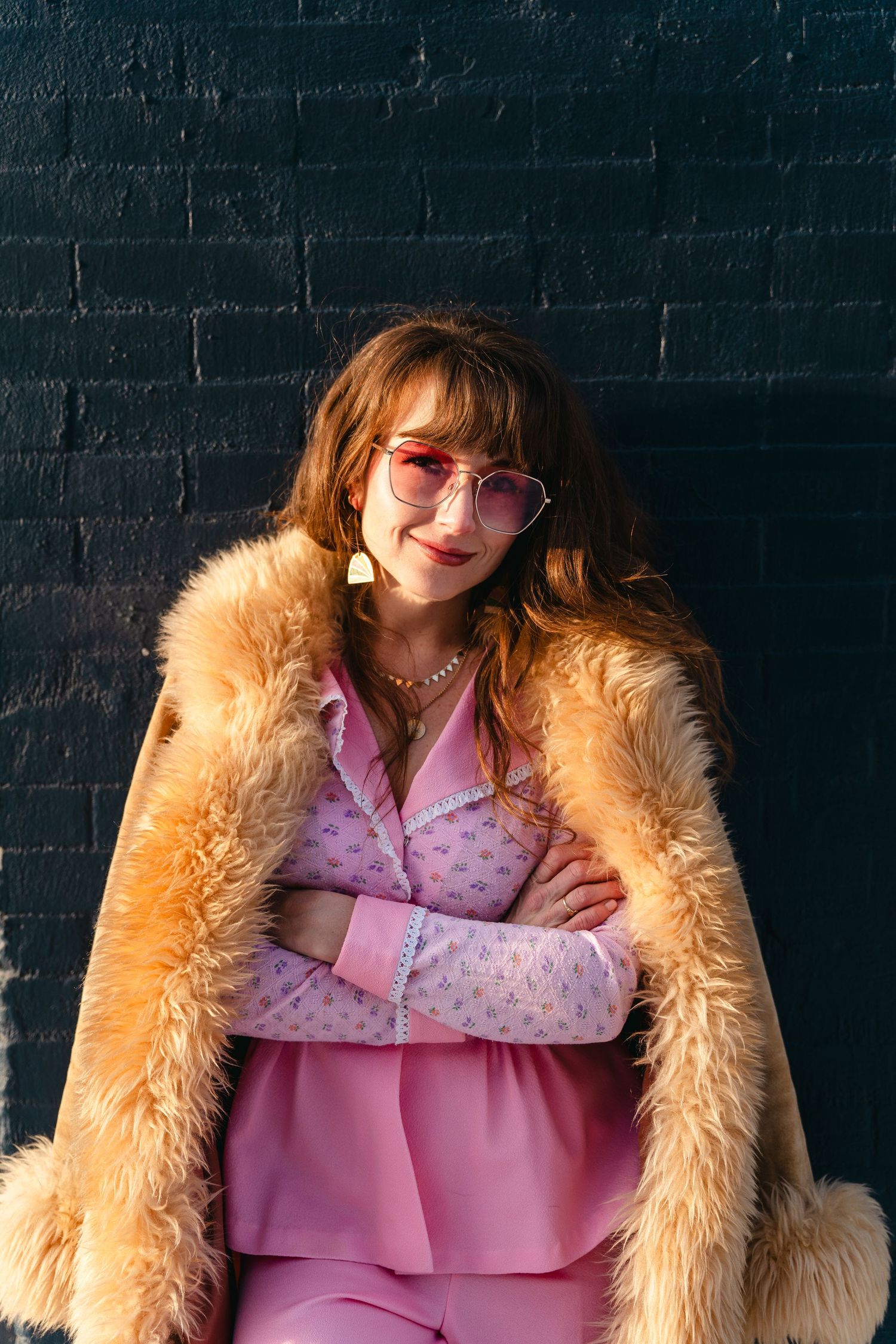 Chic retro outfit featuring pink polka dot ensemble and tan faux fur coat against dark brick wall in vintage fashion style.