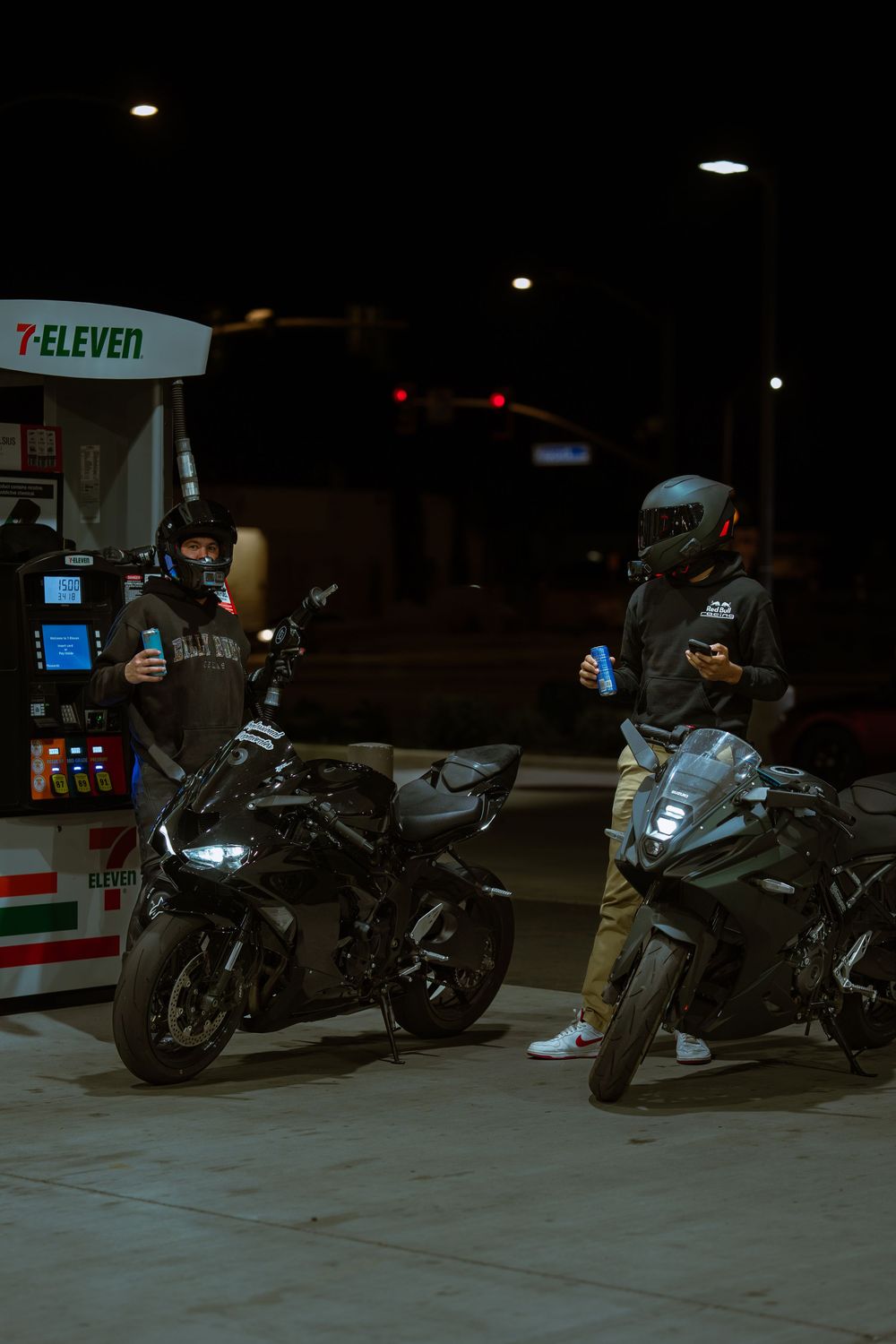 A motorcyclist in dark clothing stands next to their parked motorcycle at a gas station at night.