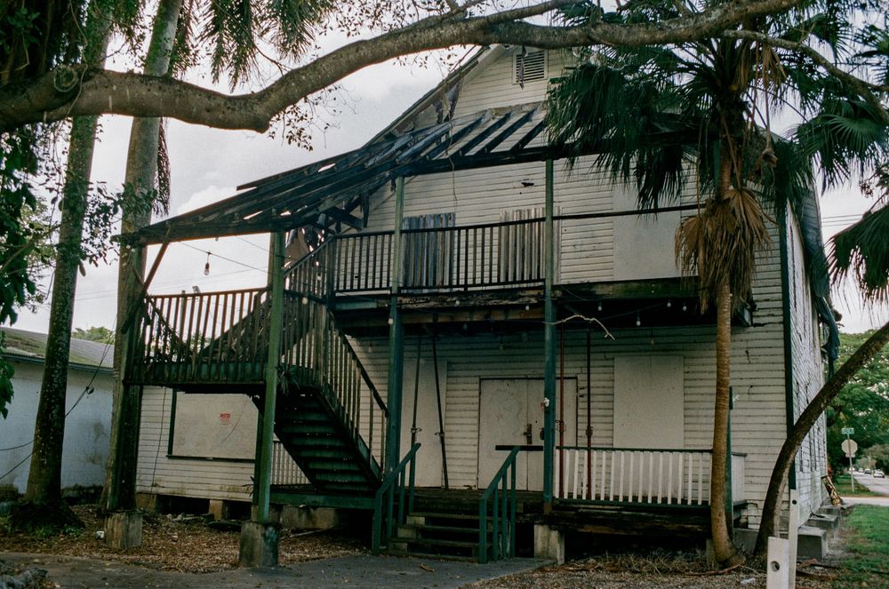 Two-story white house with exterior stairs and covered porch surrounded by tall trees.