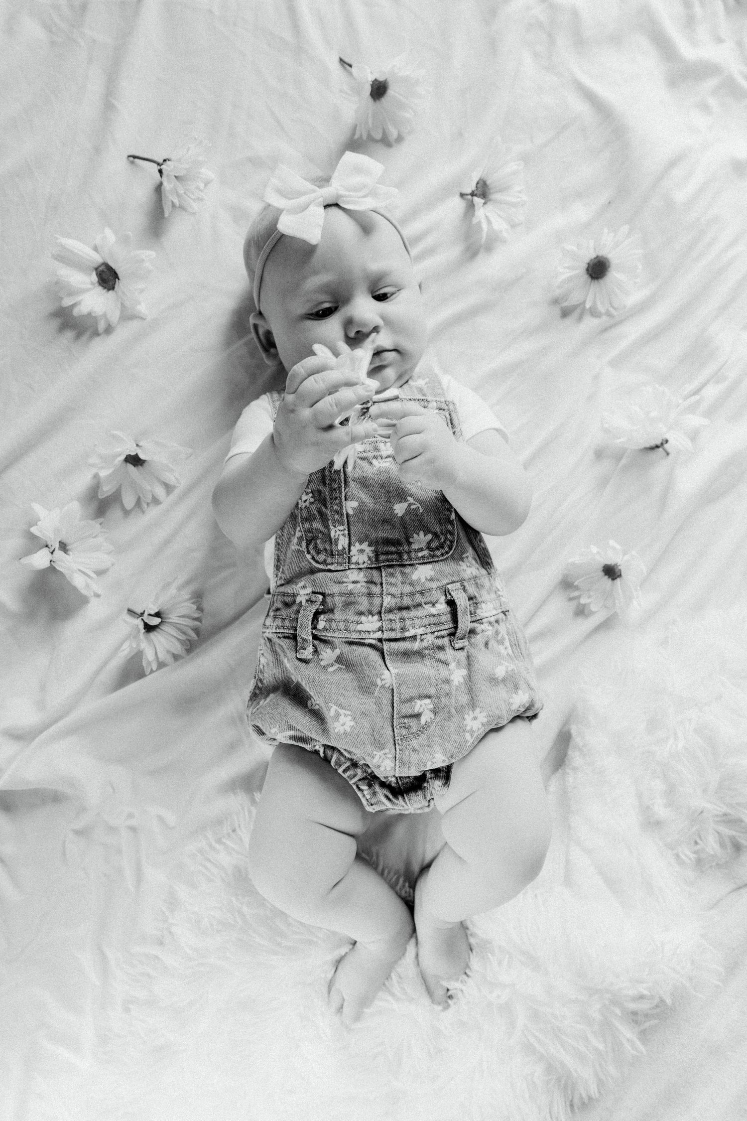Black and white photo of a baby in denim overalls lying on fabric with daisy flowers scattered around.