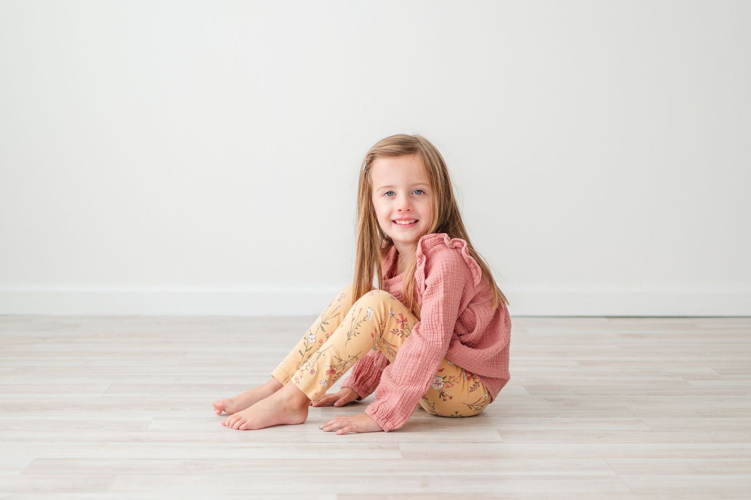 A child in pink and gold polka dot pajamas sits on a light wooden floor against a white wall.