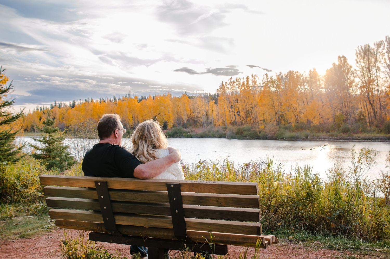A couple sits together on a wooden bench overlooking a lake surrounded by golden autumn trees at sunset.