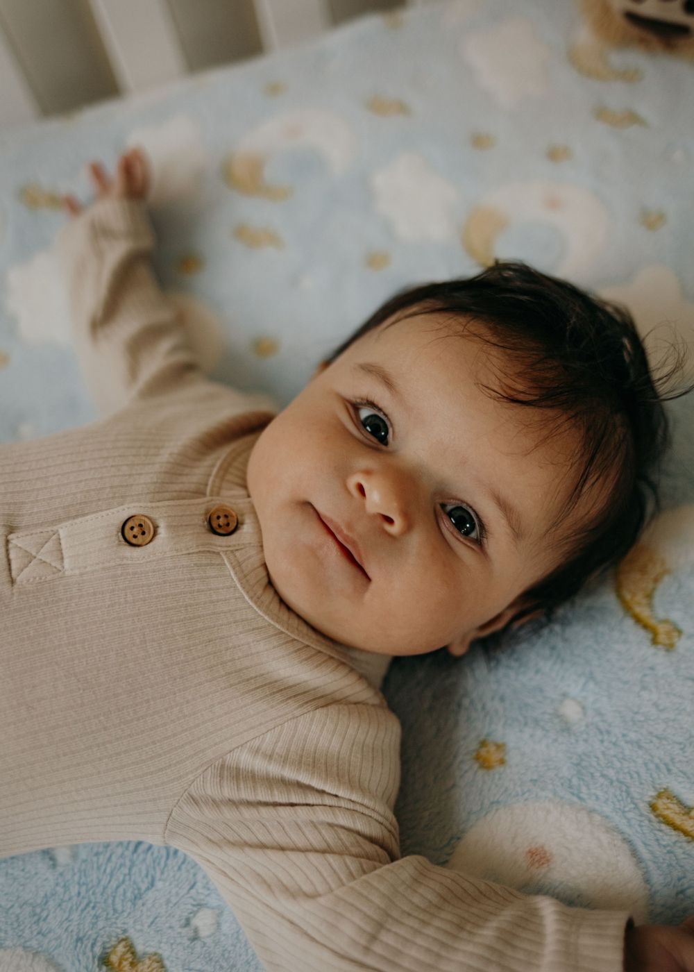 Baby wearing beige sweater lying on blue patterned bedding while looking up at camera with bright eyes.
