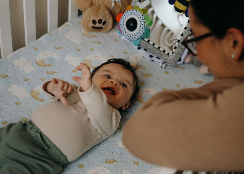 A caregiver and baby share a joyful moment together while lying on a bed with colorful bedding.