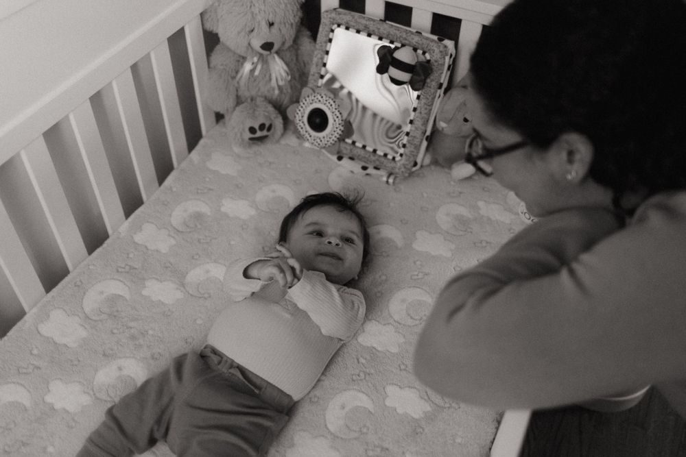 A caregiver leans over a white crib where an infant lies on a patterned mattress in black and white.