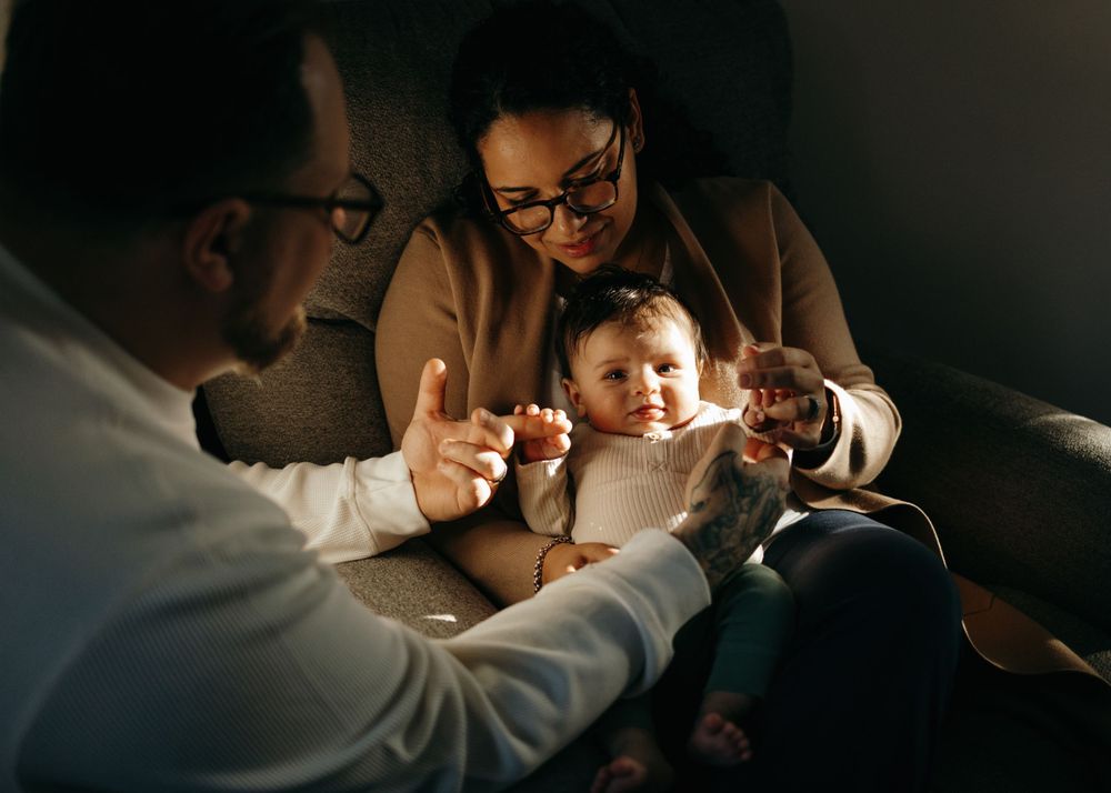 A small baby smiles while sitting with adults in a dimly lit room with warm lighting.