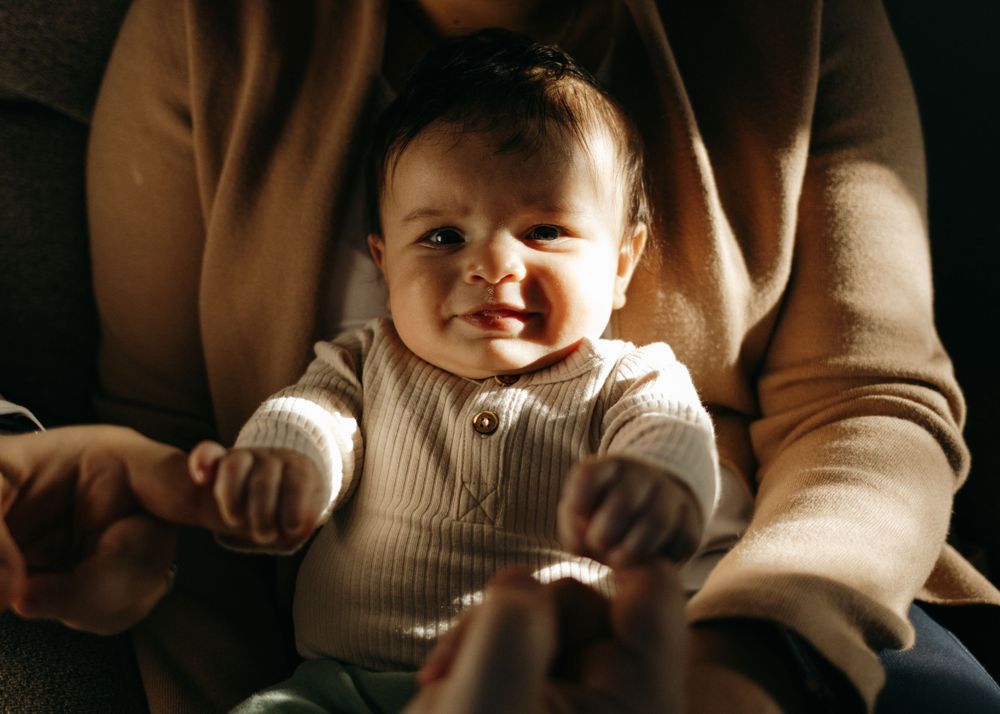 A baby wearing a white ribbed sweater smiles while being held by someone in a brown cardigan.