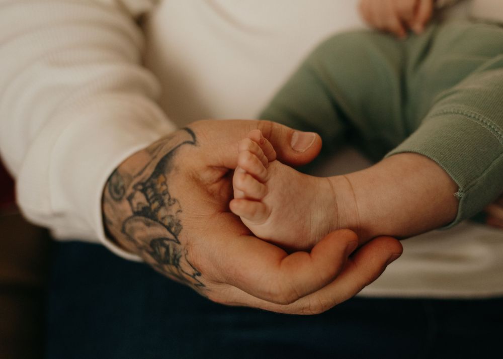 Close-up of tattooed hand gently holding a tiny baby's hand against a soft background.