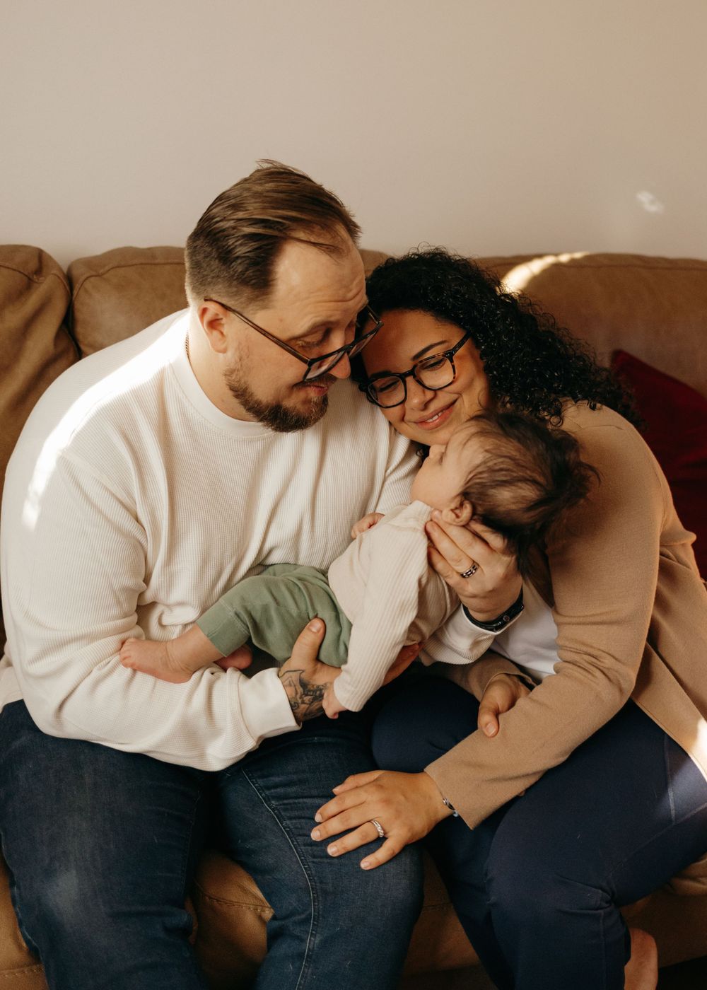 A couple in glasses cuddles their baby on a brown couch in an intimate family moment.