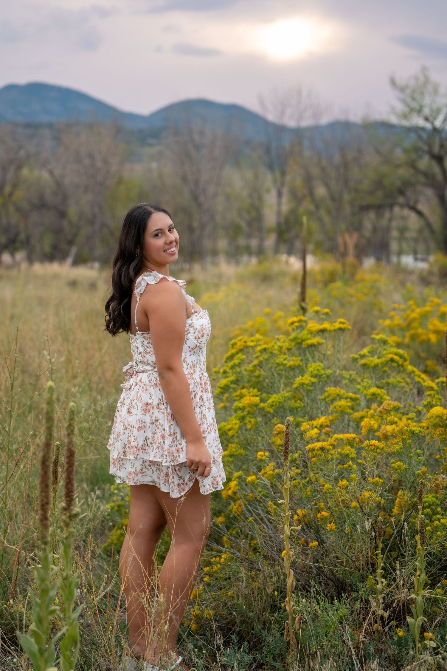 A white floral sundress stands out against yellow wildflowers and mountains in a scenic nature setting at sunset.