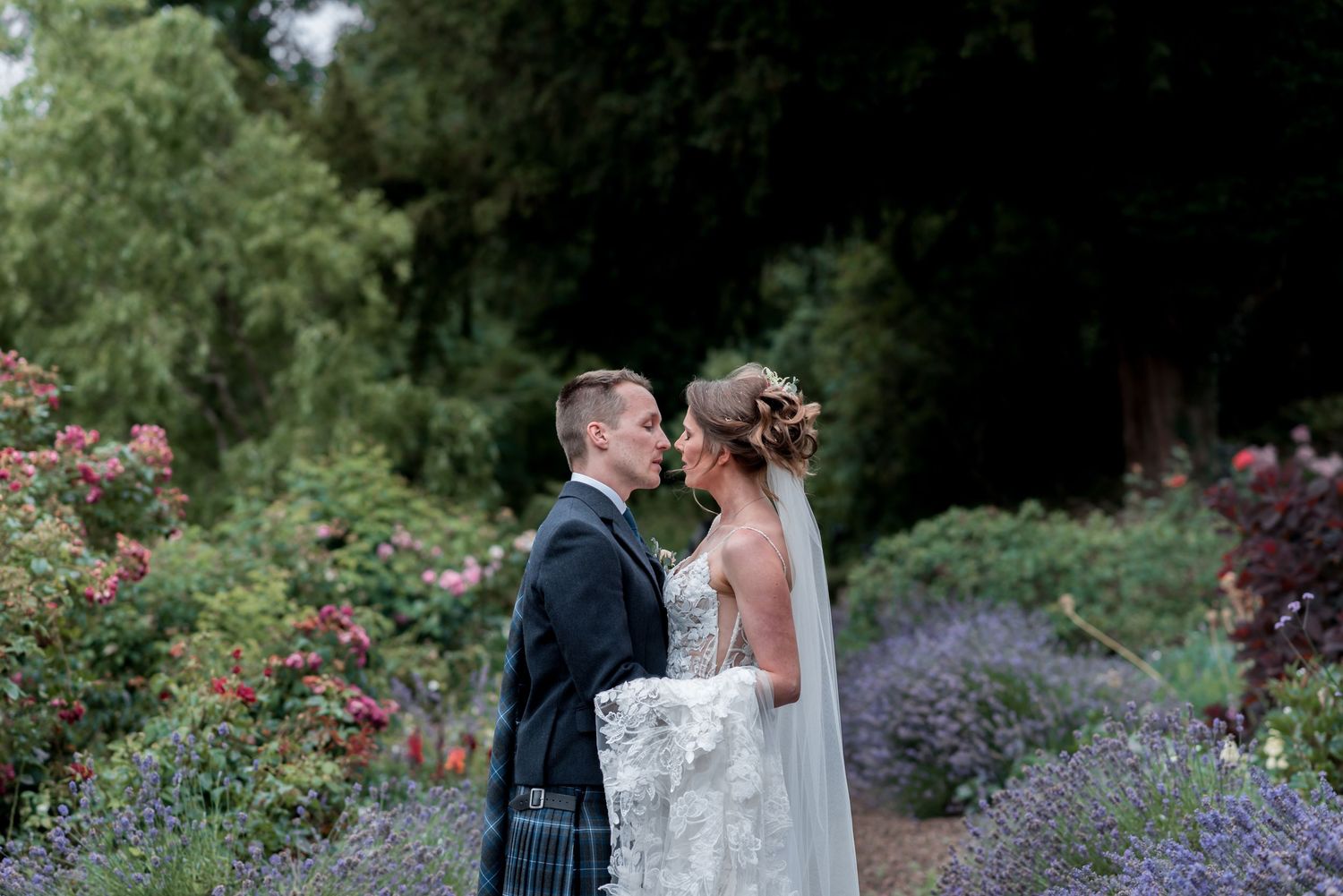 A couple shares an intimate wedding moment in a lush garden with pink roses and lavender blooms.