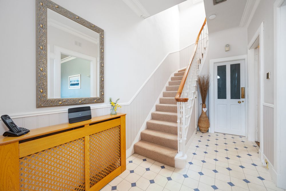 Modern hallway with white walls, carpeted stairs, decorative mirror, and a yellow radiator cover on patterned floor tiles.