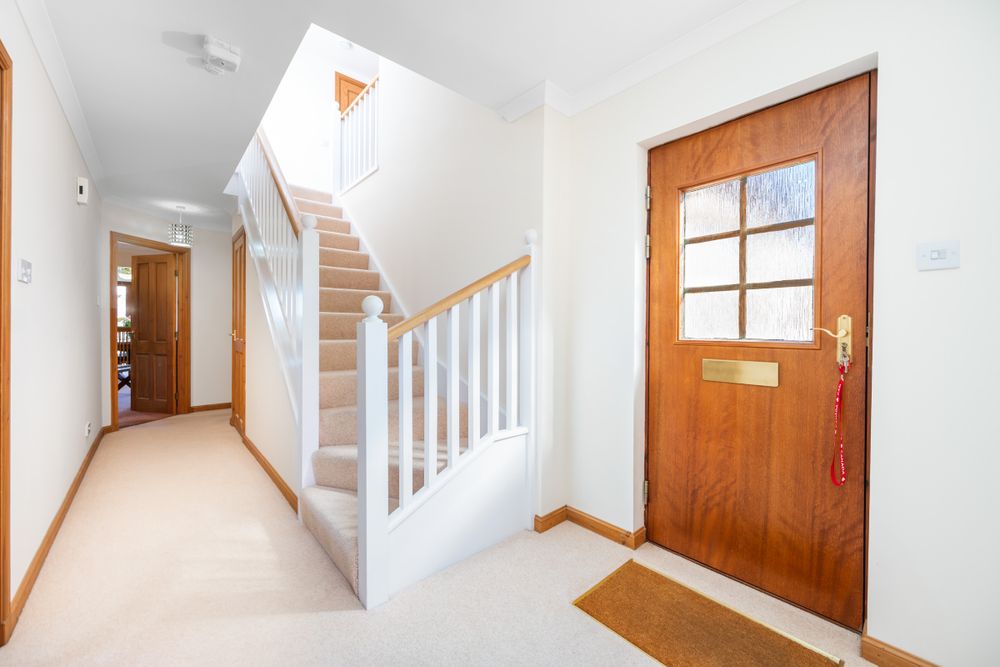 Bright interior hallway with wooden front door, staircase with white railings, and beige carpet flooring.