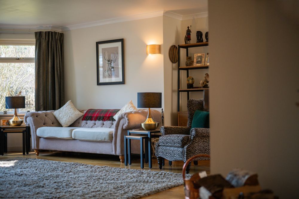 Cozy living room interior with grey sofa, patterned armchair, table lamp, and wall shelving in warm lighting.
