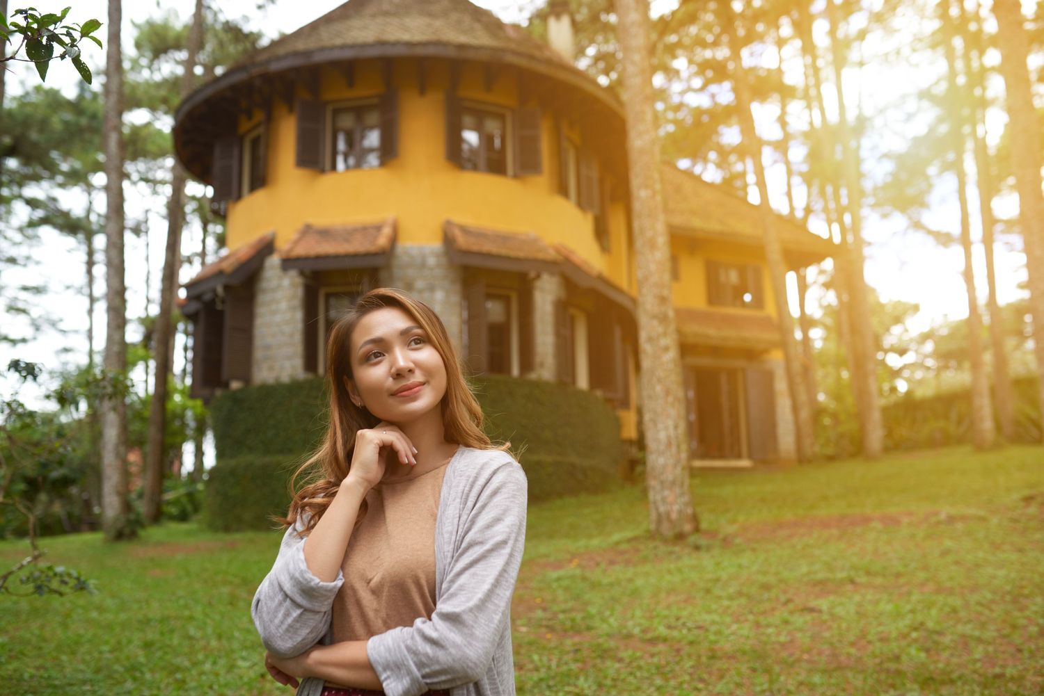 A yellow villa-style house surrounded by pine trees with sunlight filtering through.