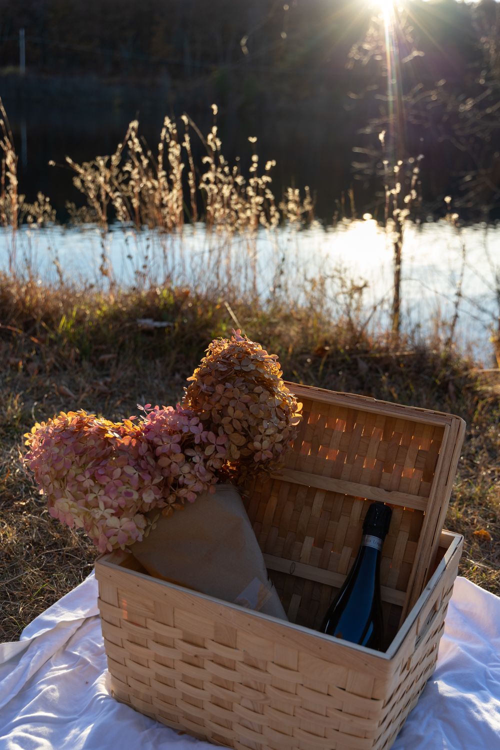 Wicker picnic basket with dried hydrangeas and wine bottle glowing in golden sunset light by lake.