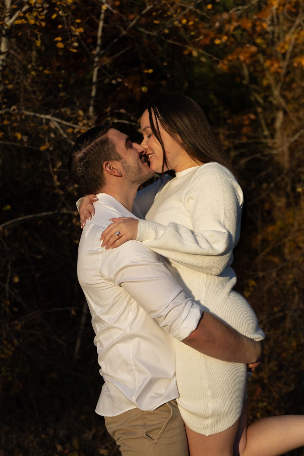 Couple in white outfits embracing and kissing surrounded by warm fall foliage.