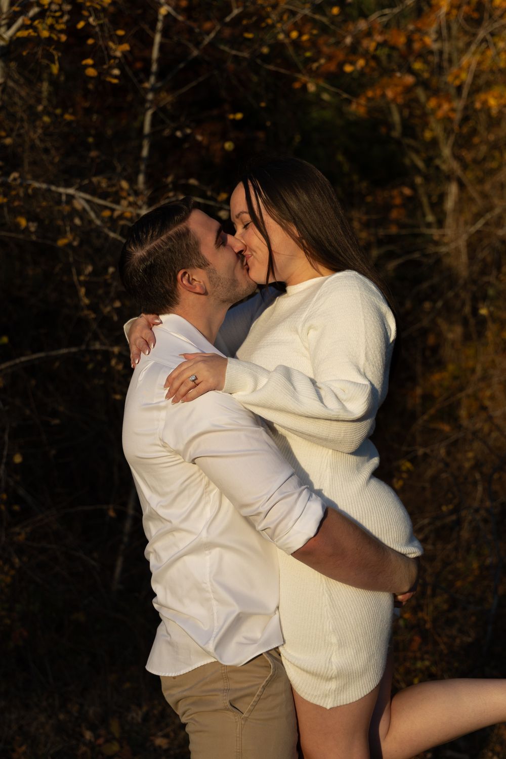 Couple in white outfits sharing a kiss surrounded by golden autumn leaves.