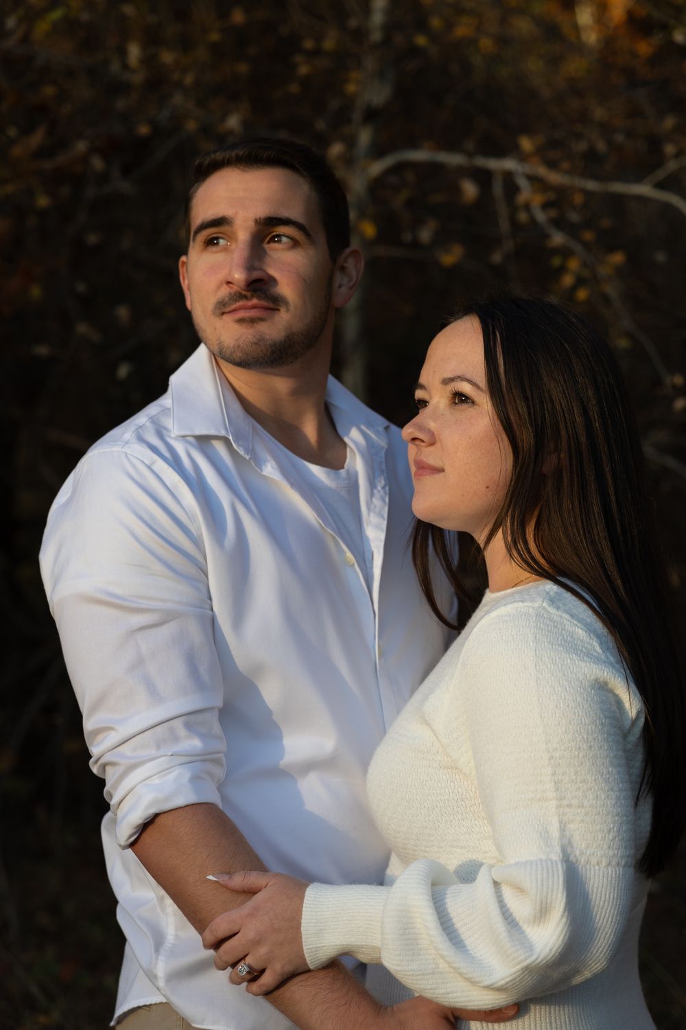 Couple holding hands and gazing upward together in golden autumn light among fall foliage.