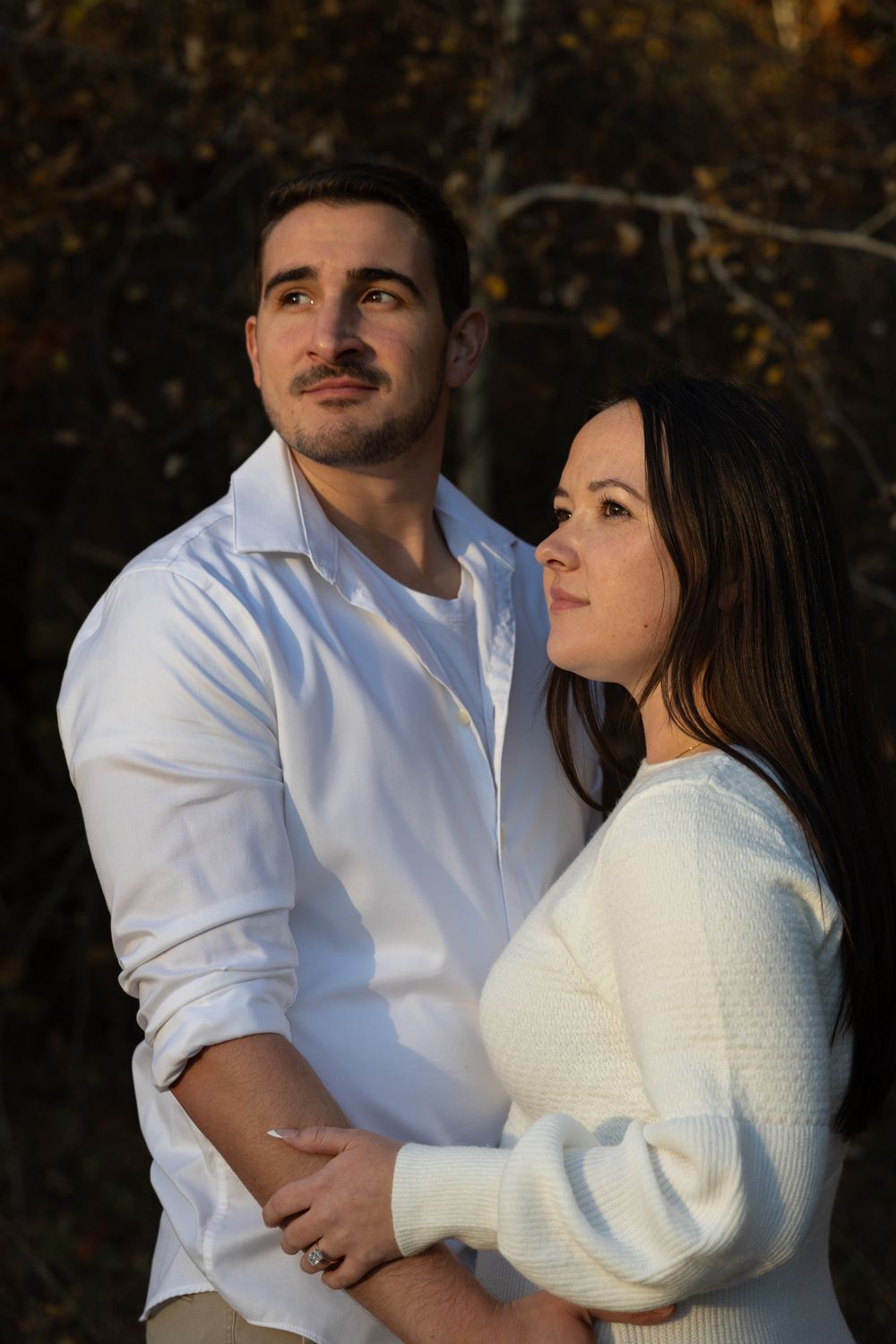 Couple embracing in warm autumn sunlight, gazing upward together among fall foliage.