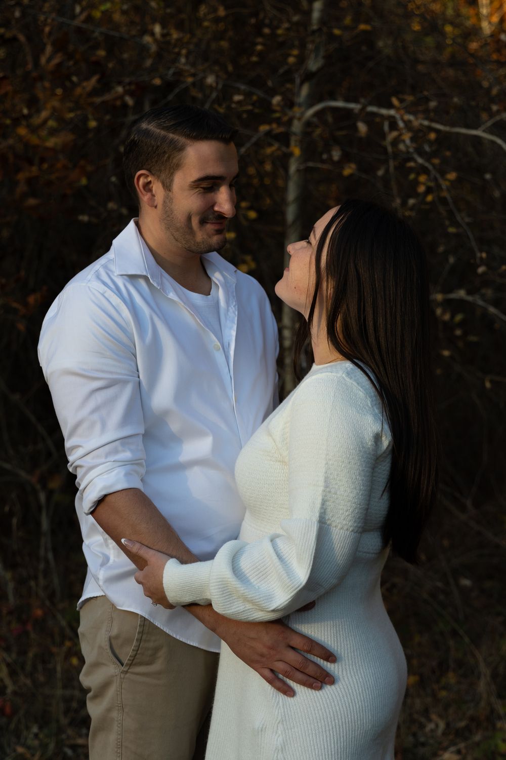 Couple in white outfits embracing romantically outdoors with autumn foliage background.