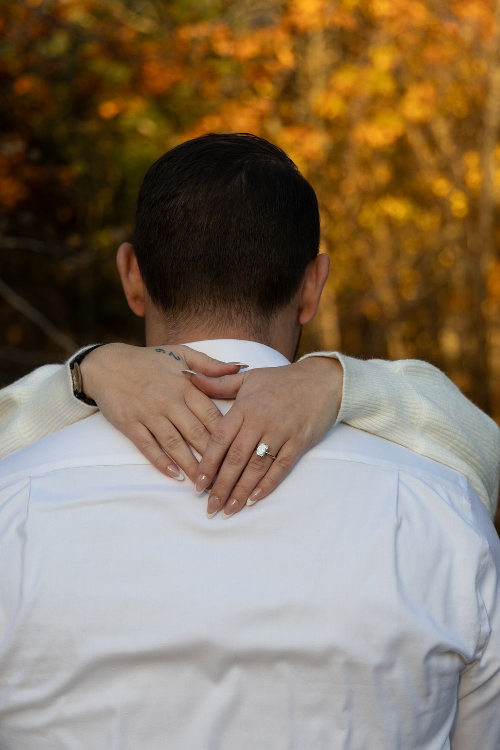 Woman's hands with diamond engagement ring clasped around partner's neck amid fall foliage.
