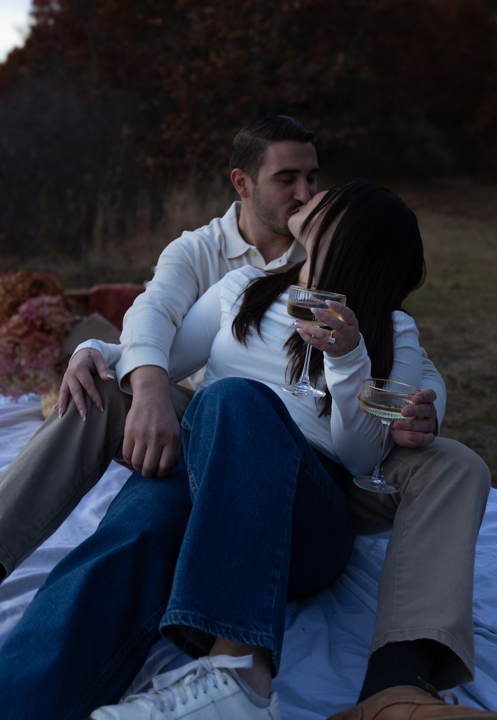 Couple sharing a kiss while holding wine glasses during a romantic autumn picnic at dusk.