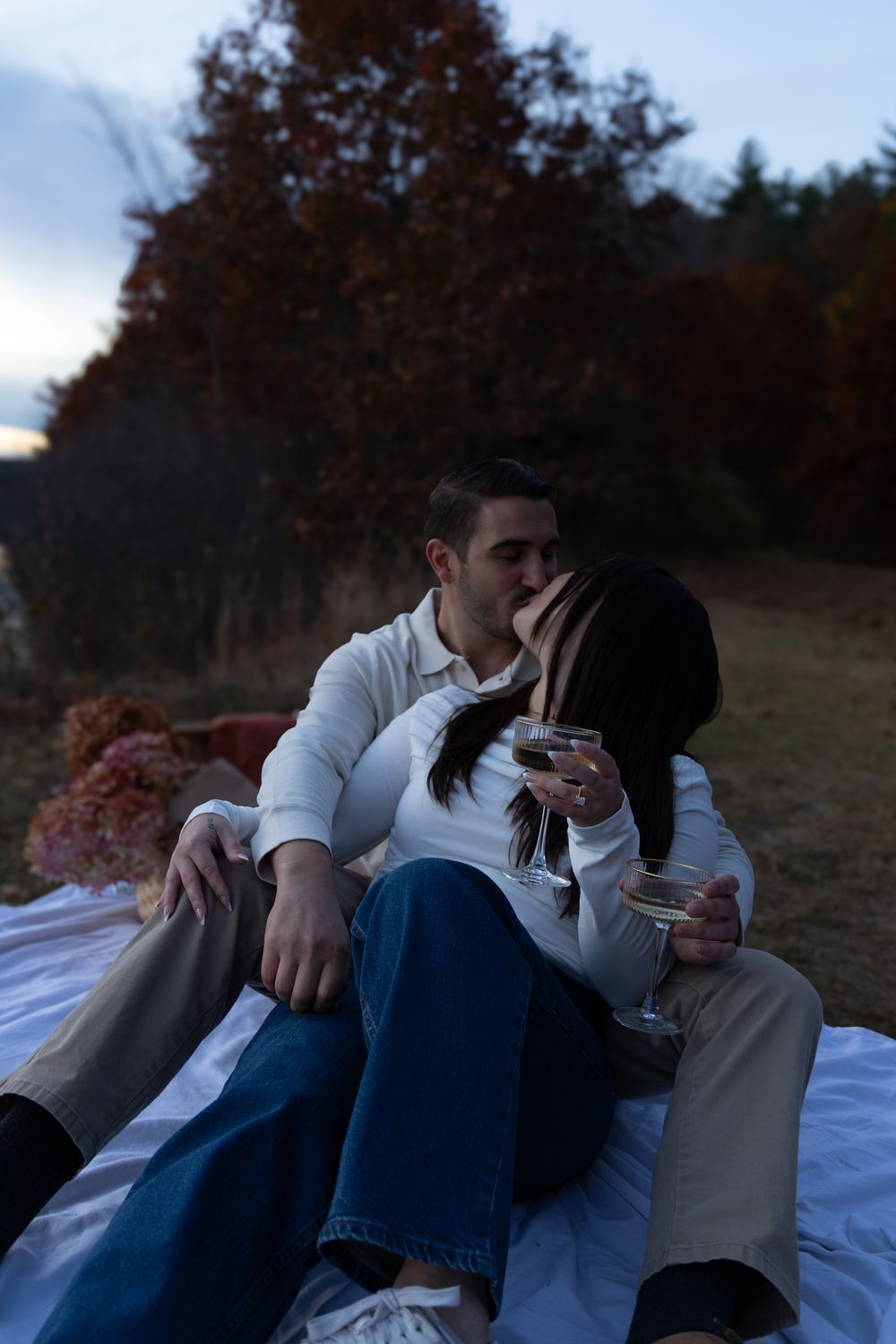 Engaged couple kissing while holding champagne glasses on a blanket at dusk outdoors.
