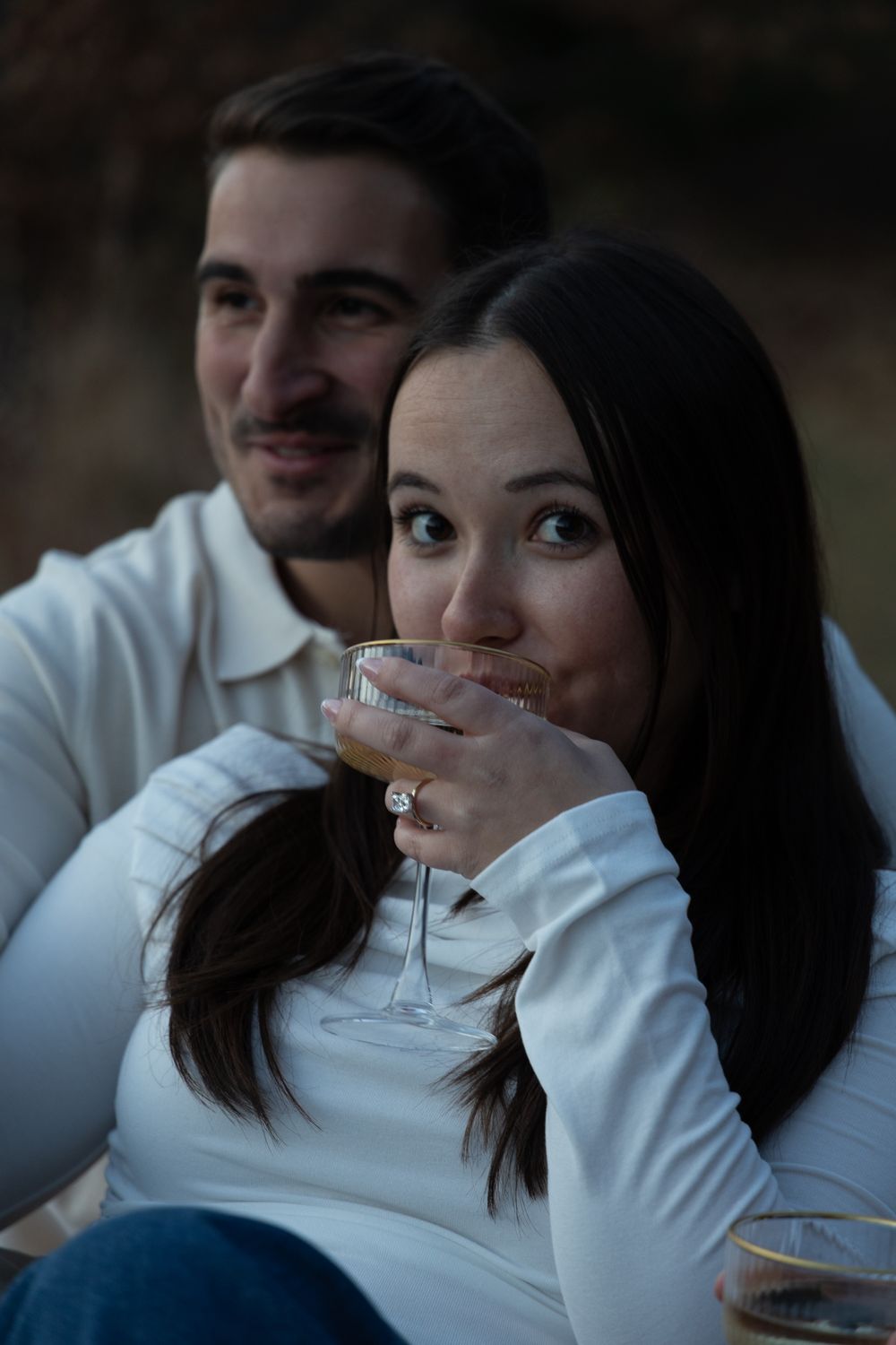 Young woman sipping champagne while her partner smiles behind her during a romantic outdoor engagement session.
